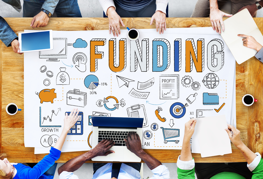 People sat at a modern desk holding a funding poster with funding related graphics like $ and graphs.